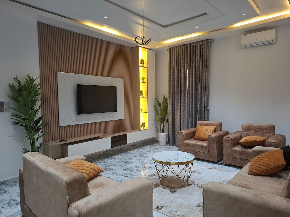 A spacious living area is featured with neutral-toned furniture, including two chairs and a sofa. A round coffee table sits at the center, surrounded by decorative plants. A mounted flat-screen television is positioned above a minimalist media unit with ambient lighting along the wall.