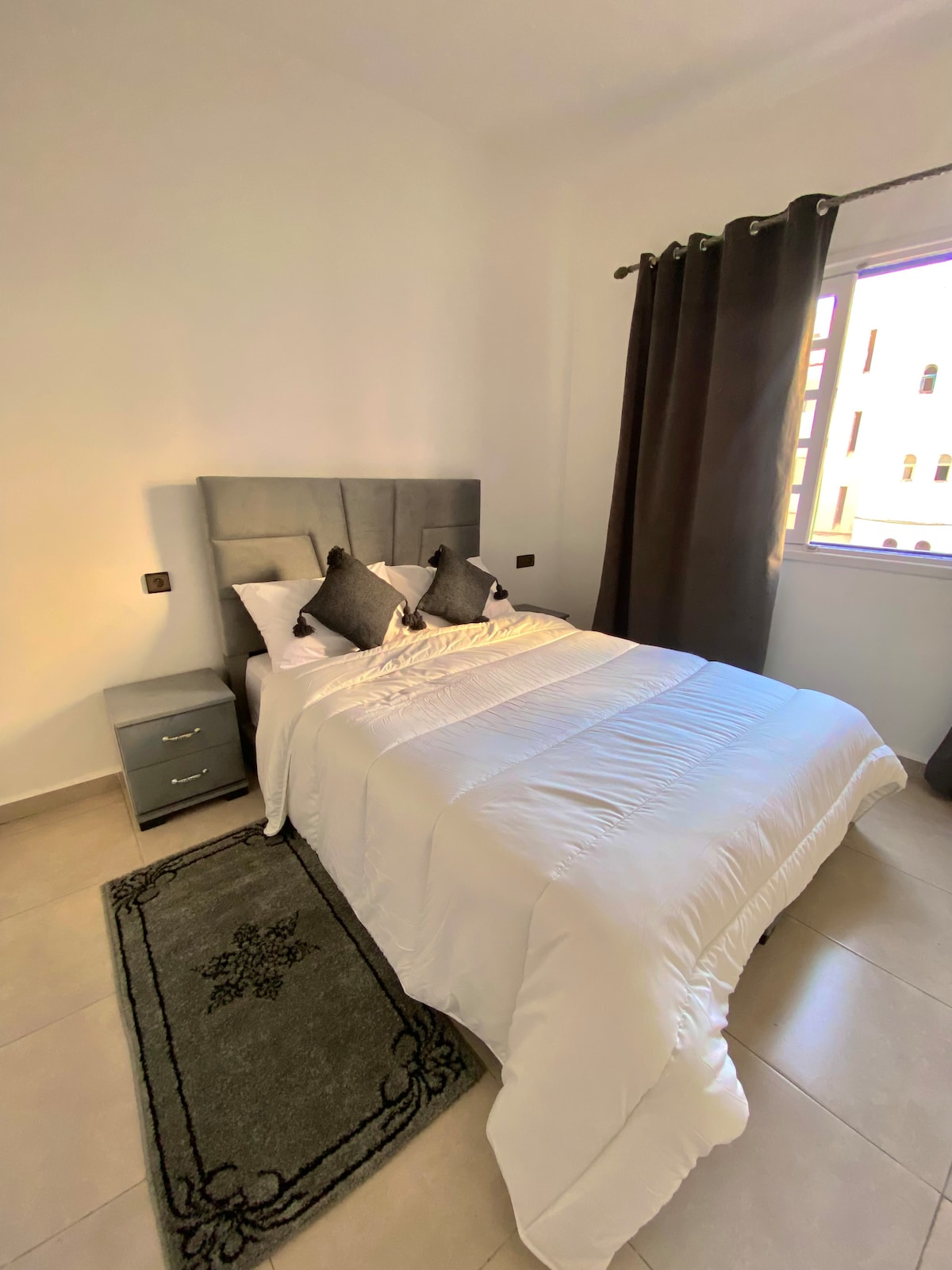 A well-lit bedroom features a spacious bed with a light-colored bedding set, positioned alongside a small bedside table. Decorative pillows are arranged neatly on the bed. A window dressed in dark curtains allows natural light to filter into the room, complementing the neutral-toned walls.