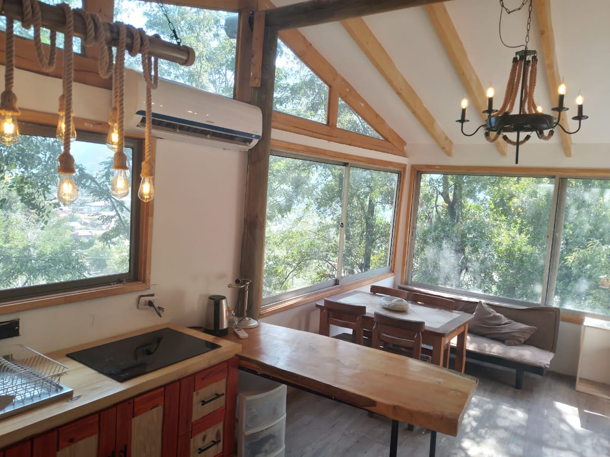 A bright kitchen area features large windows overlooking a forested landscape. Wooden beams support the high ceiling, and warm lighting hangs above a wooden dining table. A comfortable seating area with a sofa is positioned beside the windows, creating an inviting space for relaxation.