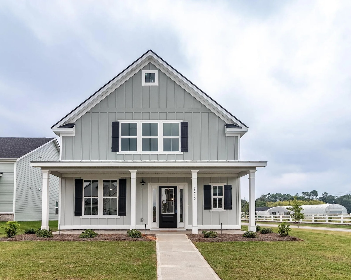 The exterior of the house features a light grey facade with white trim and black shutters. A welcoming porch is framed by tall columns, and the pathway is bordered by neatly manicured grass. The surrounding area includes a view of expansive green fields in the background.