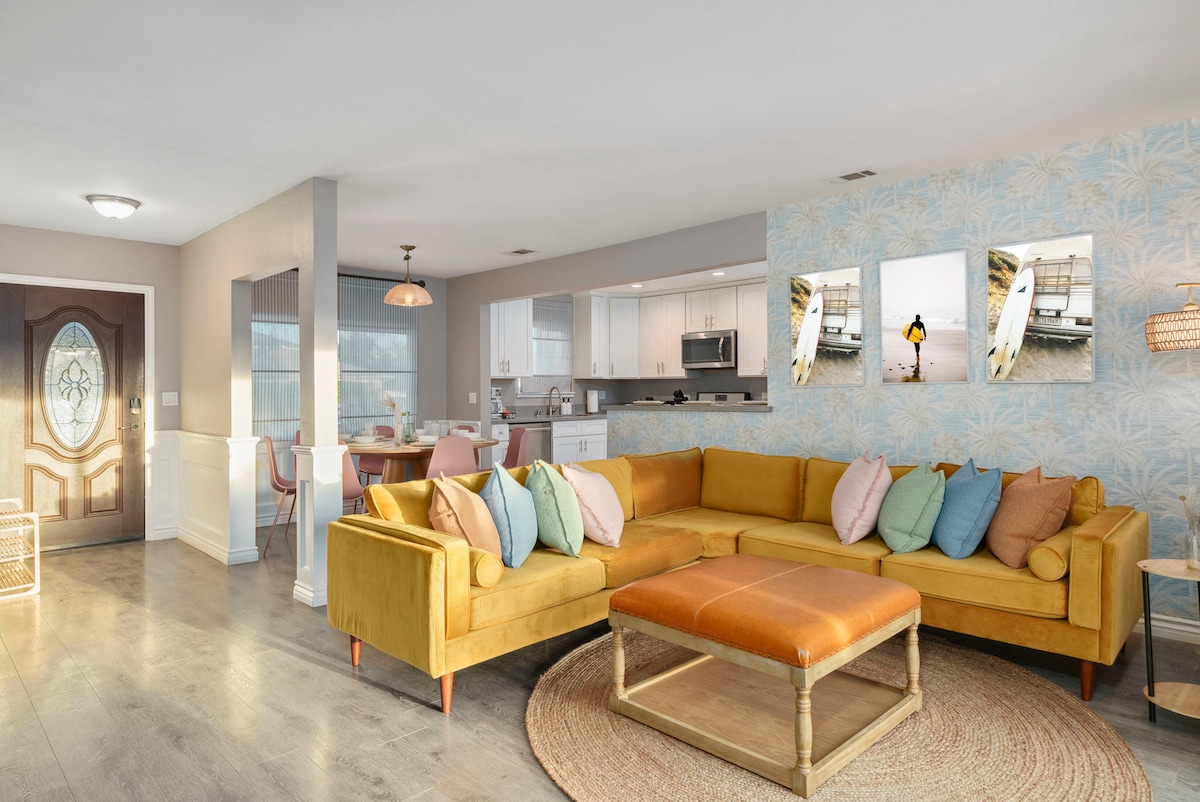 A spacious living area features a large yellow sectional sofa, adorned with multicolored cushions. Light wood flooring complements the coastal-themed wall with light blue patterns. Modern artwork of surfboards is displayed, while the open kitchen and dining area are visible in the background.