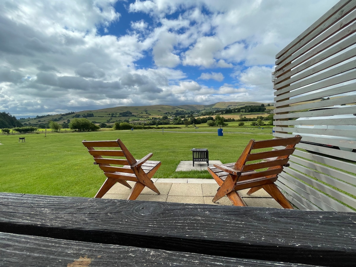 Two wooden chairs are positioned on a patio, facing a wide grassy area with expansive views of the surrounding hills. A small fire pit is visible between the chairs, and a wooden fence borders the patio, enhancing the connection to nature.