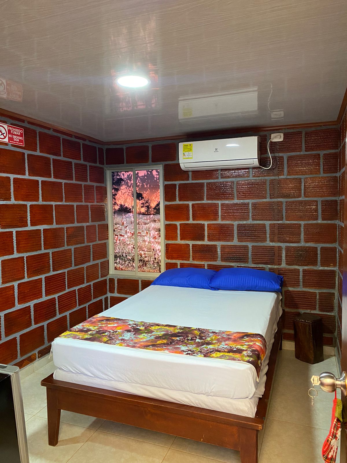 A compact room features a spacious bed with a white mattress and vibrant blue pillows. The walls are designed to resemble red bricks, creating a unique aesthetic. A window showcases a nature-themed image, while an air conditioning unit is mounted above the bed.