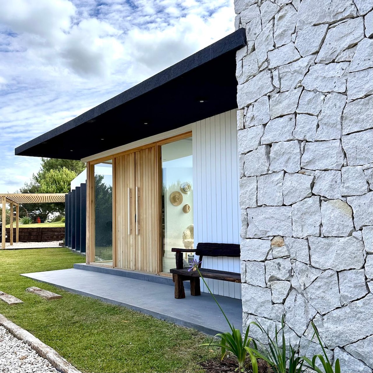 An entryway features a combination of natural stone and wooden elements. Glass double doors open to a welcoming space, complemented by a simple wooden bench and greenery in the foreground. A soft, cloudy sky adds to the tranquil outdoor setting.