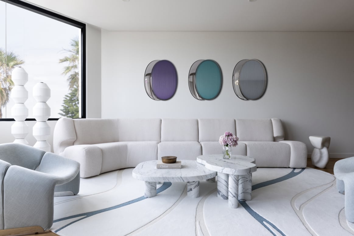 A spacious living area features a light-toned sectional sofa with a marble coffee table at its center. Circular artwork in muted colors adorns the wall, while large windows offer views of palm trees. Light pastel chairs are positioned to enhance conversation.