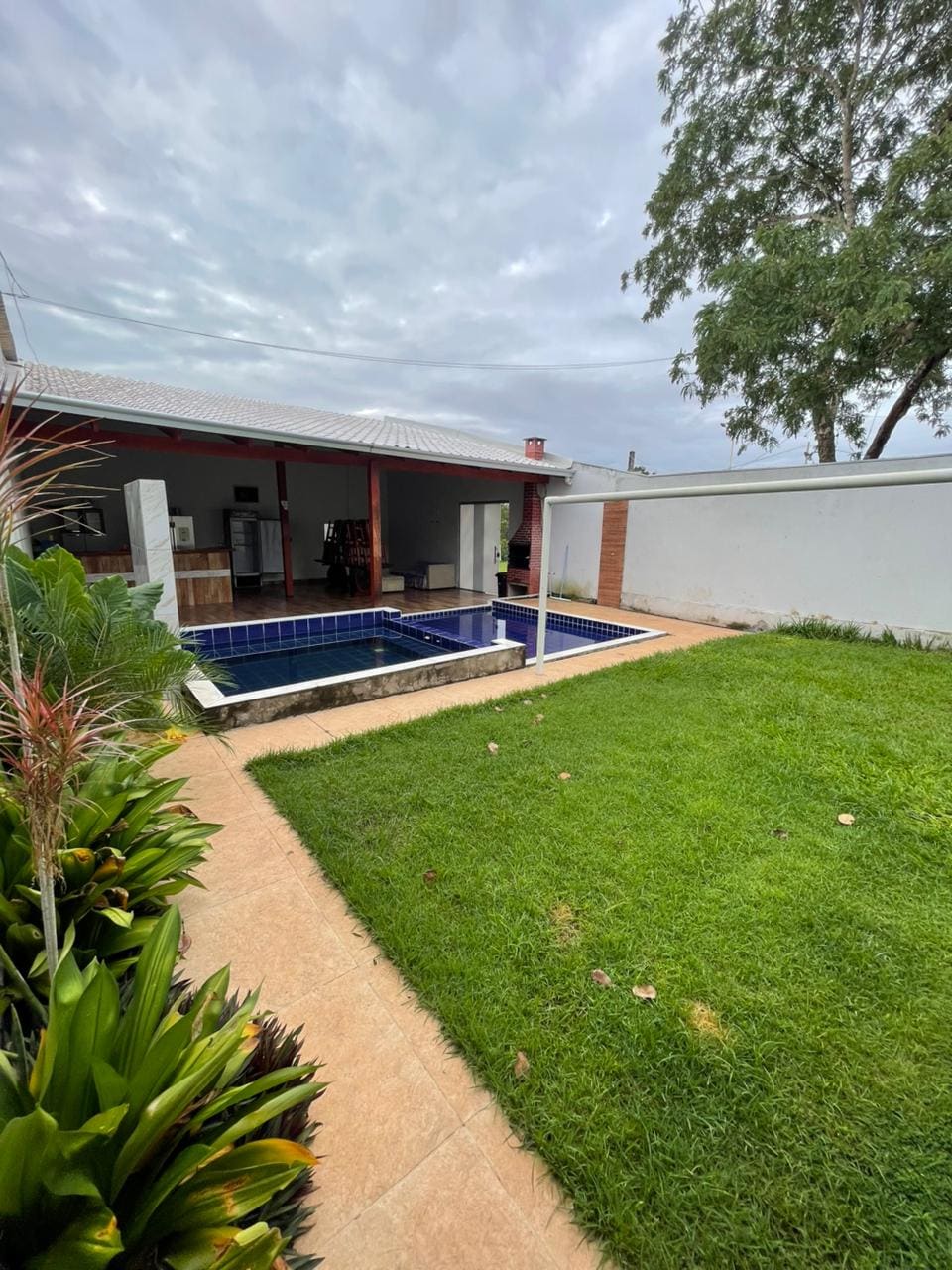 A tranquil outdoor space features a pool surrounded by manicured grass. The area is complemented by lush plants along the walkway, providing a serene setting. A covered patio area with a kitchen is visible in the background, creating a seamless flow between indoor and outdoor living.