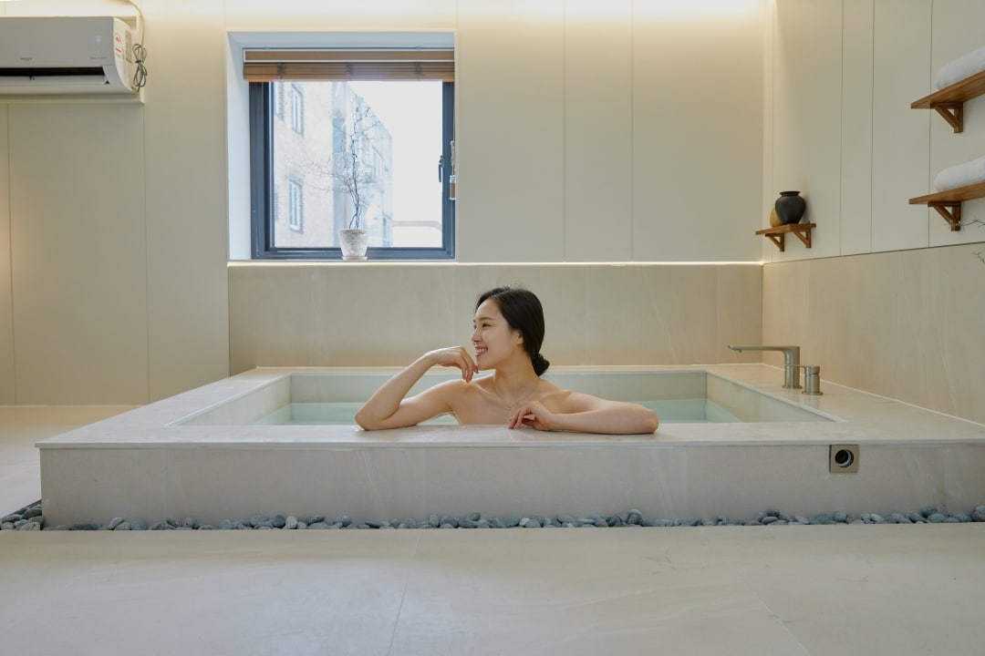 A spacious indoor spa room features a large, rectangular soaking tub surrounded by smooth stone accents. Natural light filters through a window with a wooden blind, illuminating the serene atmosphere. Minimalist decor and neutral tones create a calming environment.