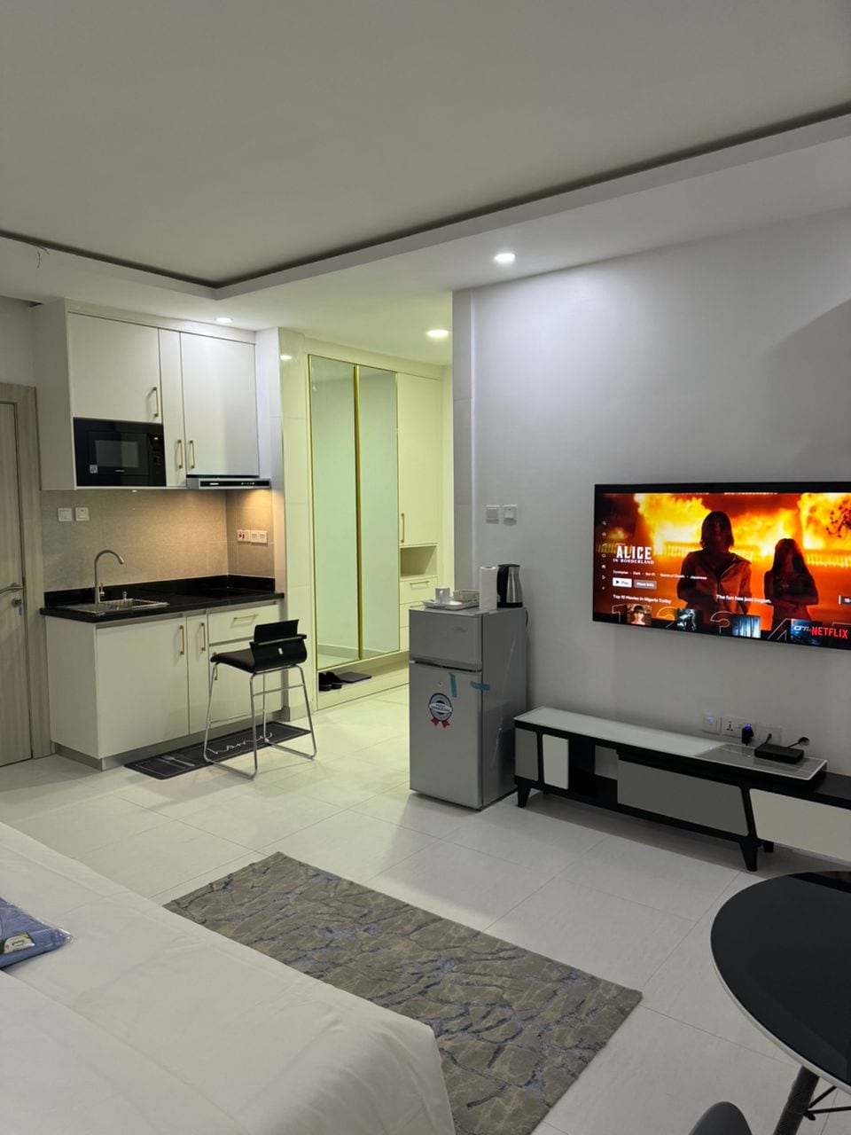 A modern studio apartment is presented, featuring a compact kitchenette with sleek cabinetry and a dining area. A flat-screen television is mounted on the wall, opposite a cozy bed adorned with a blue throw pillow. Bright lighting enhances the clean, minimalist design.