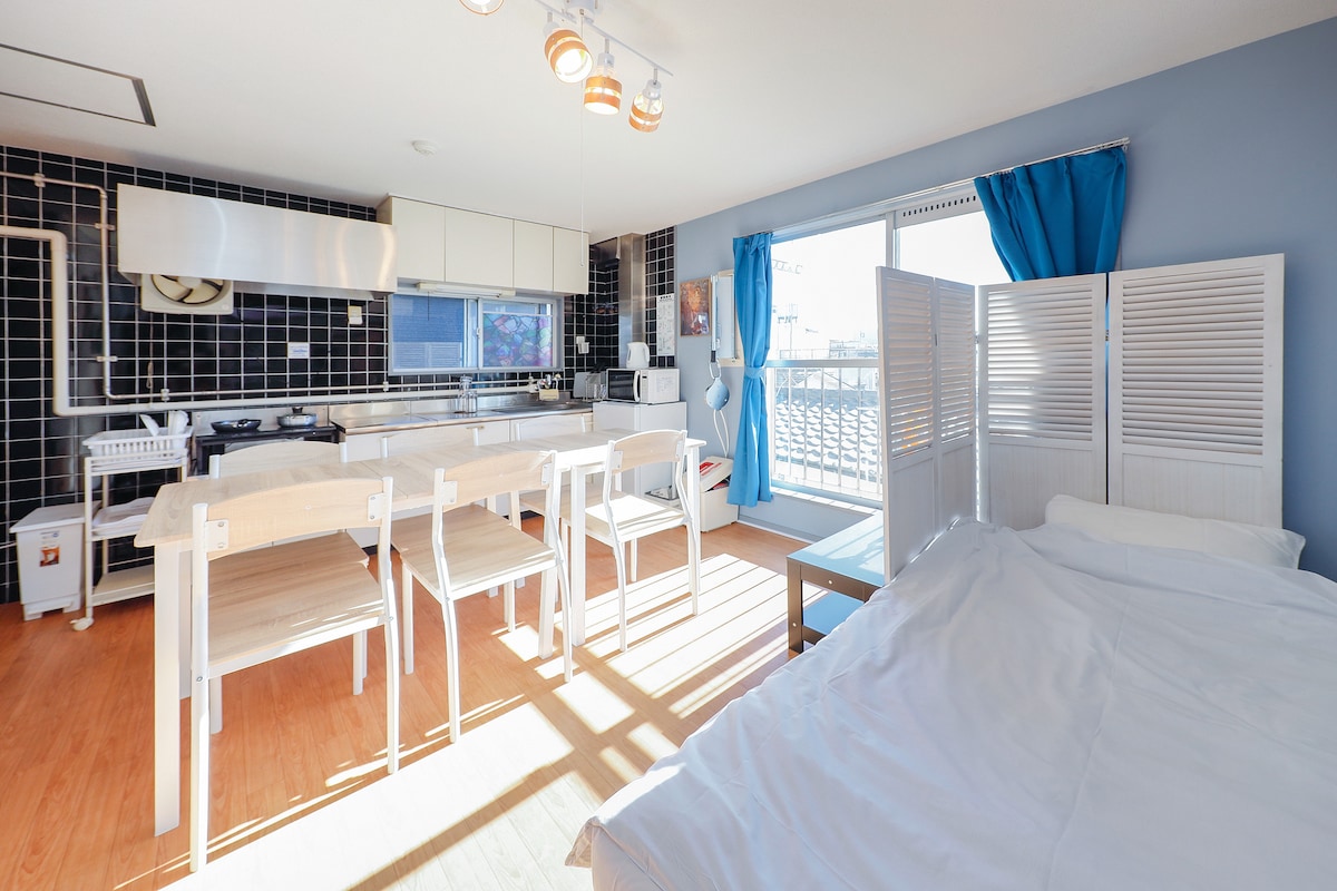 A bright open space featuring a kitchen area with modern appliances, including a two-burner stove and a refrigerator. A dining table with four chairs is positioned near the large window, allowing natural light to fill the room. A partition separates the sleeping area, which includes a single bed.