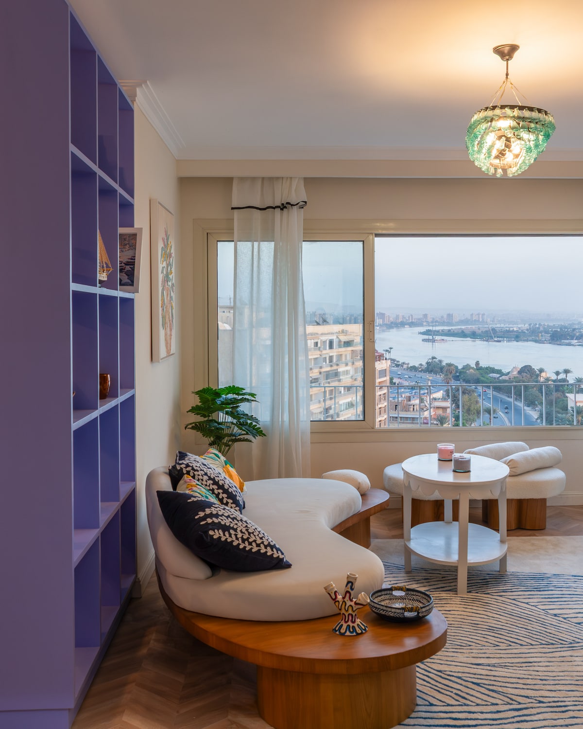 A stylish living room features a cozy curved sofa adorned with decorative cushions. A small coffee table sits nearby, complemented by a patterned area rug. Large windows provide expansive views of the river and cityscape, while a colorful chandelier adds a touch of charm.