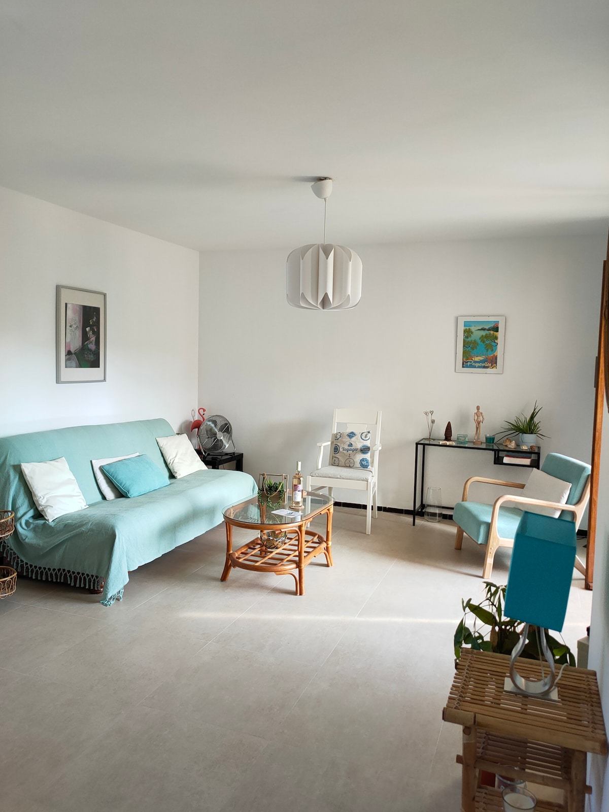 A bright living area features a teal sofa, two accent chairs, and a small glass coffee table with decorative items. Sunlight illuminates the room, enhancing a clean and airy feel. A side table displays a small plant and art piece, contributing to a relaxed atmosphere.