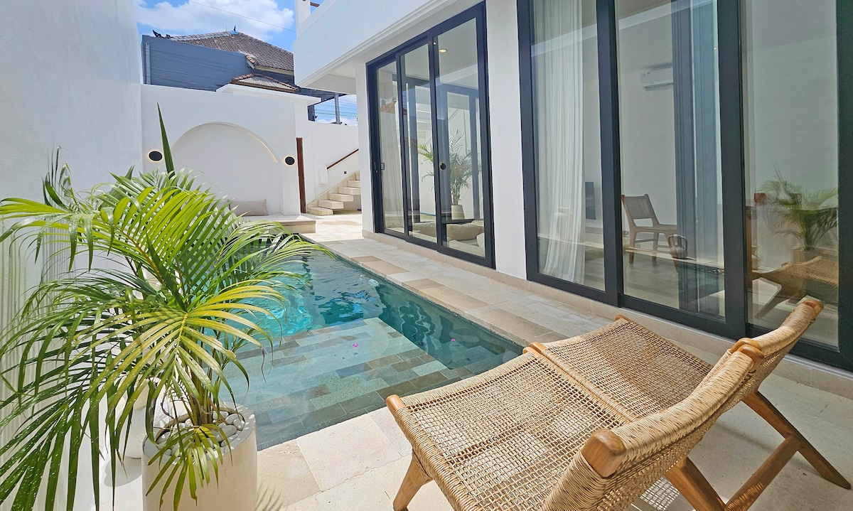 A calm private pool area features clear water surrounded by sleek tile and elegant glass doors. Two loungers are positioned nearby, and a potted plant adds a touch of greenery. Natural light fills the space, enhancing the serene atmosphere.
