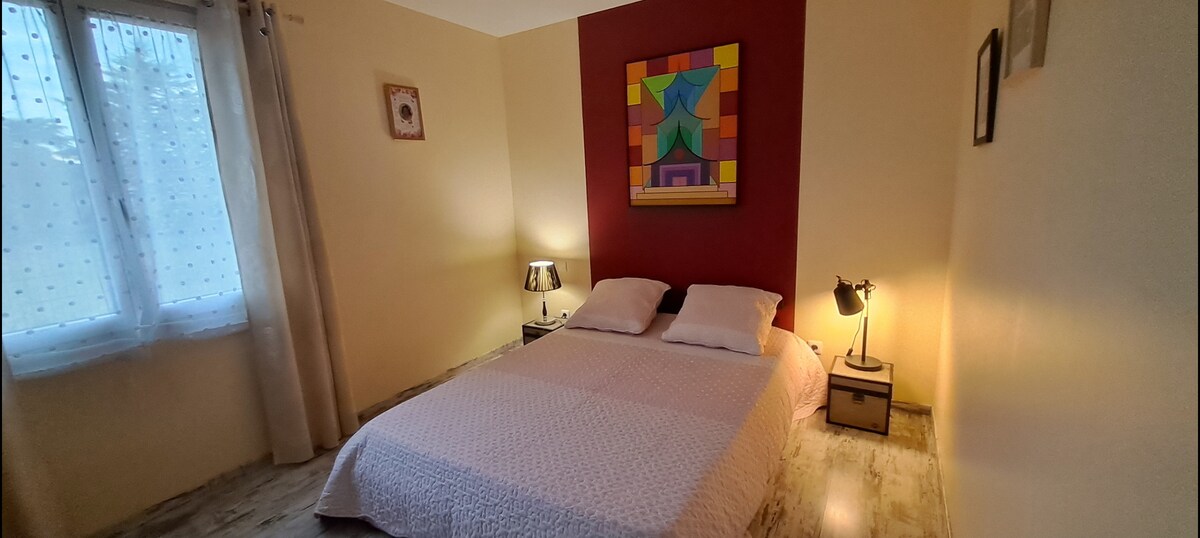 A cozy bedroom is presented with a spacious bed covered in a light quilt. A colorful wall art piece is displayed above the bed, complemented by warm bedside lamps. Natural light enters through a window, while soft curtains frame the scene.