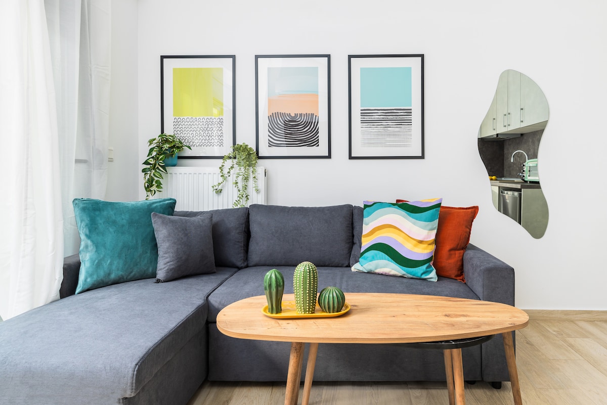 A spacious living area is highlighted by a gray sectional sofa adorned with colorful cushions. Above the sofa, three contemporary framed artworks in soft tones are displayed. A round wooden coffee table holds a trio of decorative cacti, while a modern kitchen area is visible in the background.