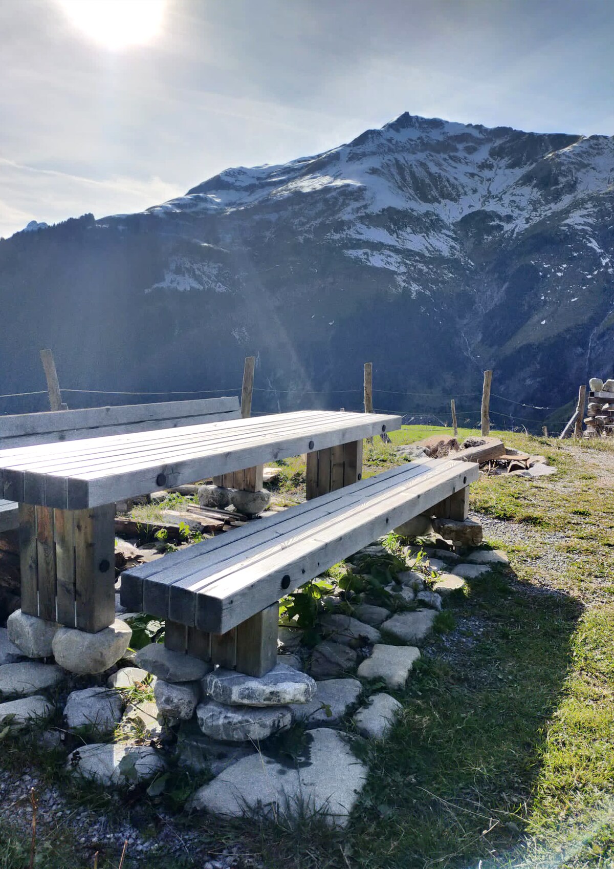 A wooden picnic table with attached benches is positioned on a grassy area, surrounded by rocky stones. Majestic snow-capped mountains loom in the background under a clear sky, hinting at the natural beauty of the location.