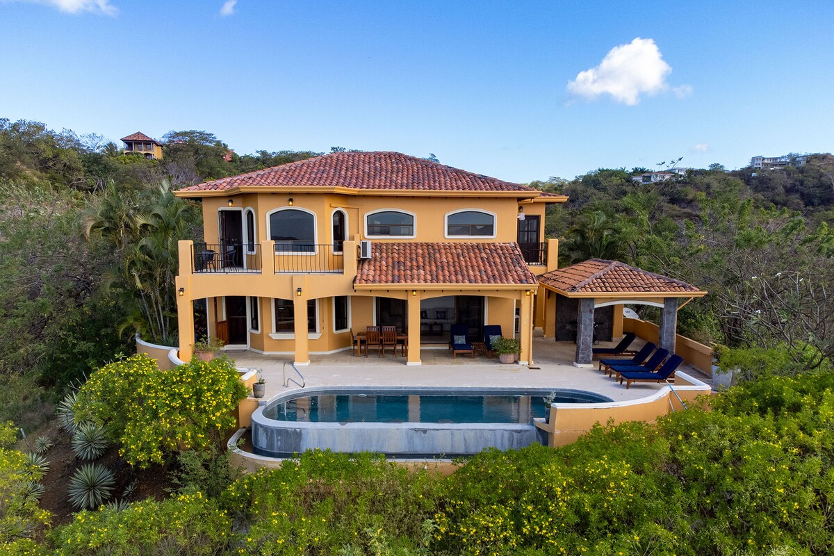 The exterior of a large villa is surrounded by lush tropical foliage, featuring a tiled roof and multiple balconies. An infinity pool extends toward the horizon, accompanied by outdoor seating and a shaded lounge area, creating a picturesque outdoor living space.