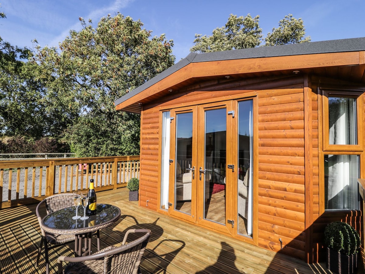 The exterior of the lodge features a wooden facade with large glass doors that open to a decked area. A round table with two chairs and a bottle of champagne is set on the decking, surrounded by trees that provide enhancing privacy.