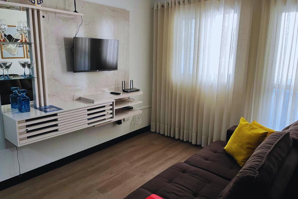 A cozy living area is presented, featuring a mounted television above a neutral-colored media console. Natural light filters through sheer curtains, illuminating the space. A comfortable sofa with a relaxed cushion setup and a yellow throw pillow adds a pop of color.