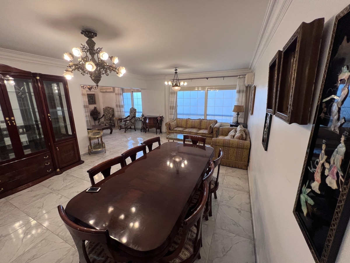 A spacious dining area is presented with a large wooden table surrounded by matching chairs. Large windows allow natural light to fill the room, highlighting the elegant chandelier above. The space features classic furniture pieces and tasteful decor that reflects a blend of modern and traditional styles.