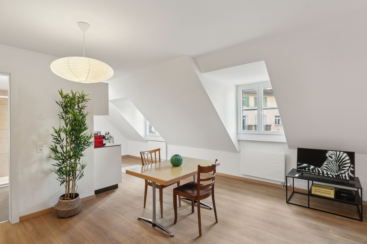 A bright dining area is featured with a wooden table and two matching chairs. A green plant adds a touch of nature to the space. A television is positioned against the wall, and large windows provide ample natural light, highlighting the angular ceiling.