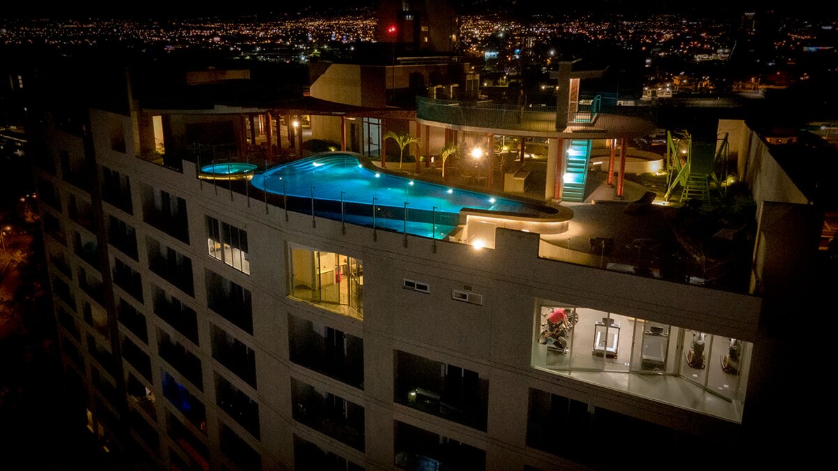 The elevated rooftop area is illuminated at night, showcasing a heated infinity pool surrounded by lounge seating and palm trees. The city lights create a vibrant backdrop, while areas of the building reveal indoor facilities through expansive windows.