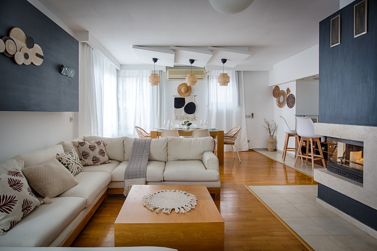 A contemporary living area features a large, L-shaped sectional sofa adorned with decorative pillows. A wooden coffee table with a circular placemat sits at its center. Natural light filters through sheer curtains, illuminating the adjacent dining area with a wooden table and stylish chairs.