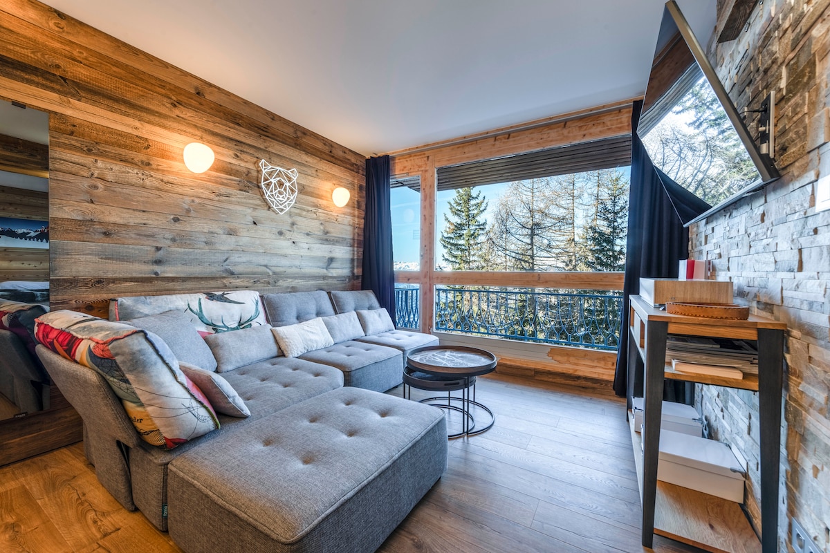 A cozy living area features a spacious gray sectional sofa adorned with decorative cushions. Natural wood accents enhance the warmth of the space, while large windows provide ample natural light and an outdoor view of trees. A small table and entertainment unit are also visible.