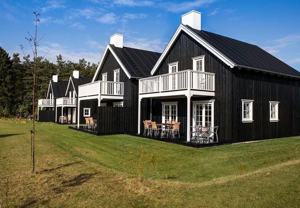 Two adjacent wooden houses are showcased in a modern architectural style, featuring black exteriors with white accents. Each house displays a balcony and outdoor seating in the front yard, surrounded by well-maintained green grass and a clear blue sky.