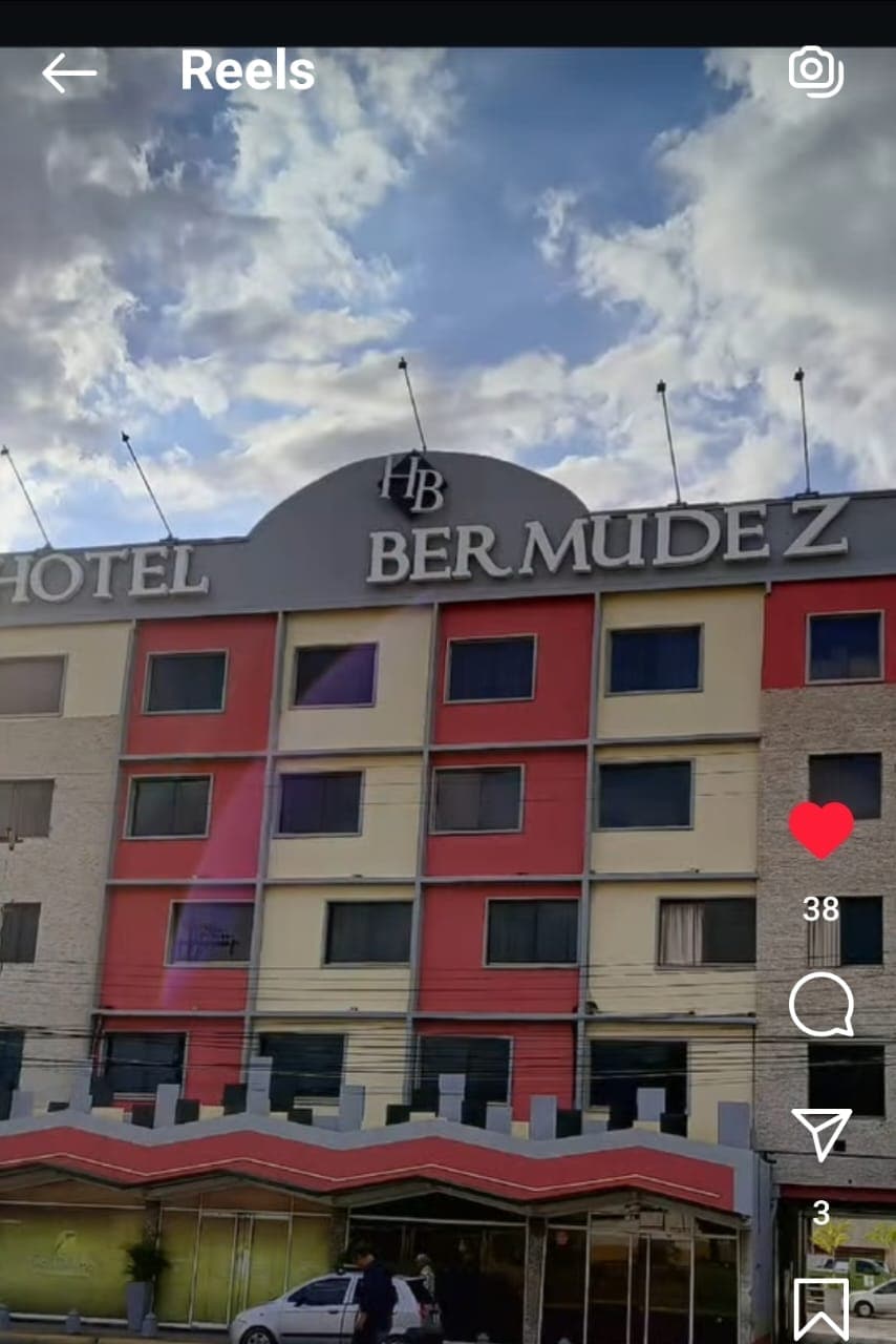 The exterior of a hotel building is displayed, featuring a modern design with a combination of red, yellow, and beige panels. The hotel's name, 'Bermudez,' is prominently displayed on the rooftop, beneath a partly cloudy sky. A covered entrance is visible at the front.
