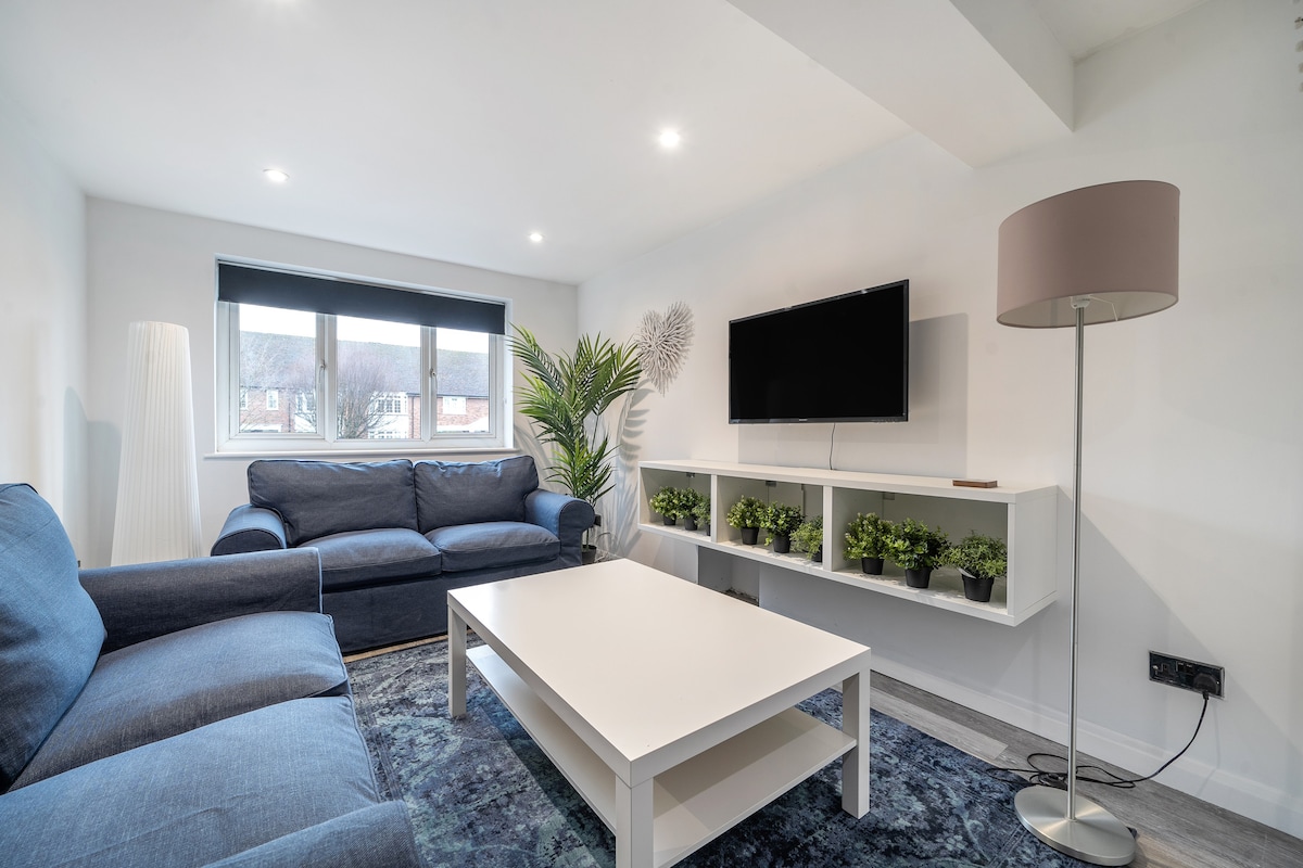 A modern living area is presented with two blue sofas and a white coffee table at the center. A flat-screen television is mounted on the wall, next to a shelf that holds small potted plants. Natural light enters through large windows, complemented by soft lighting fixtures.