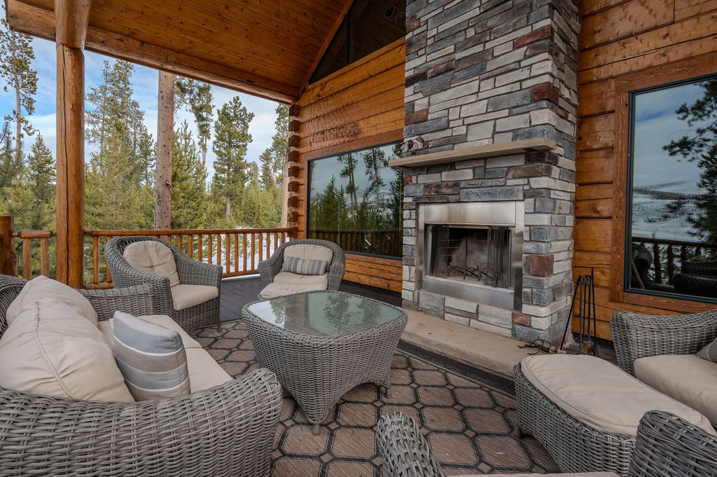 A spacious covered deck is adorned with comfortable seating arranged around a textured coffee table. A stone fireplace creates a focal point, surrounded by large windows that offer views of the surrounding trees and mountains.