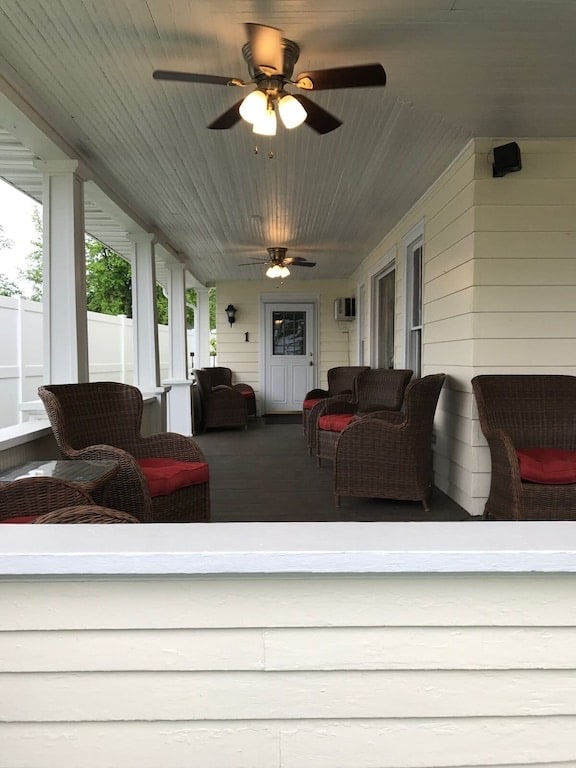 A covered porch features several wicker chairs with red cushions, arranged for seating. Ceiling fans provide ventilation, while the entrance door to the suite is visible in the background. Light-colored walls and a wooden floor create an inviting space.