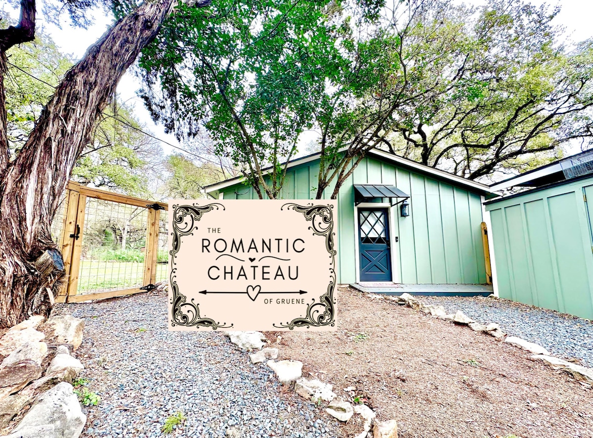 The entrance to the Romantic Chateau is framed by trees, with a gravel path leading to a welcoming wooden gate. A decorative sign featuring the name of the chateau is displayed prominently, complementing the rustic yet charming aesthetic of the property.