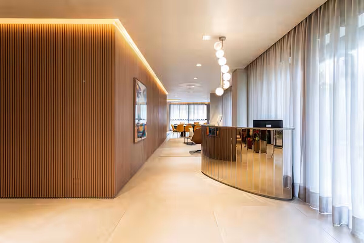 A sleek reception area is featured with wooden paneling and soft lighting. A reflective curved counter is positioned prominently, complemented by large windows draped in sheer curtains. The space leads to a well-lit seating area with modern furniture in the background.