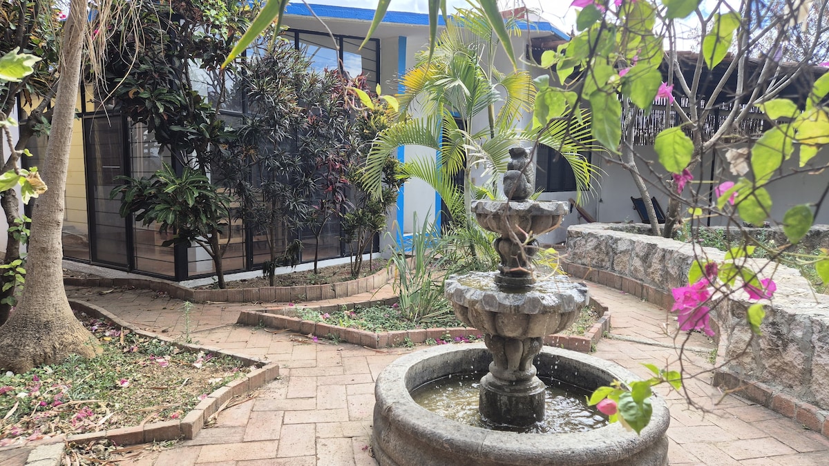 A serene garden space features a classic stone fountain at the center, surrounded by a circular stone path. Lush greenery from various plants and flowering vines provides a tranquil atmosphere, while pink blooms add subtle color to the landscape.