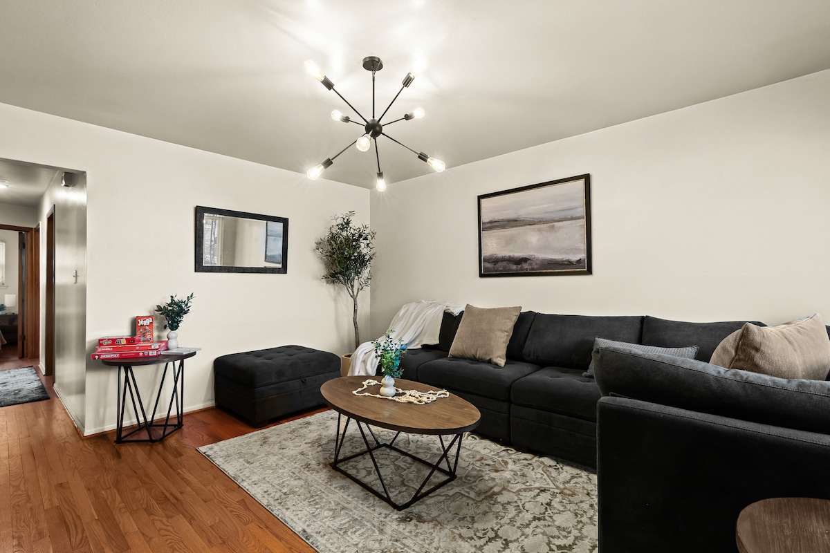 A modern living area is presented with a sectional sofa adorned with neutral-toned cushions. A round coffee table sits center stage, showcasing decorative items. Artwork is displayed on the walls, and ambient lighting is provided by a distinctive ceiling fixture.
