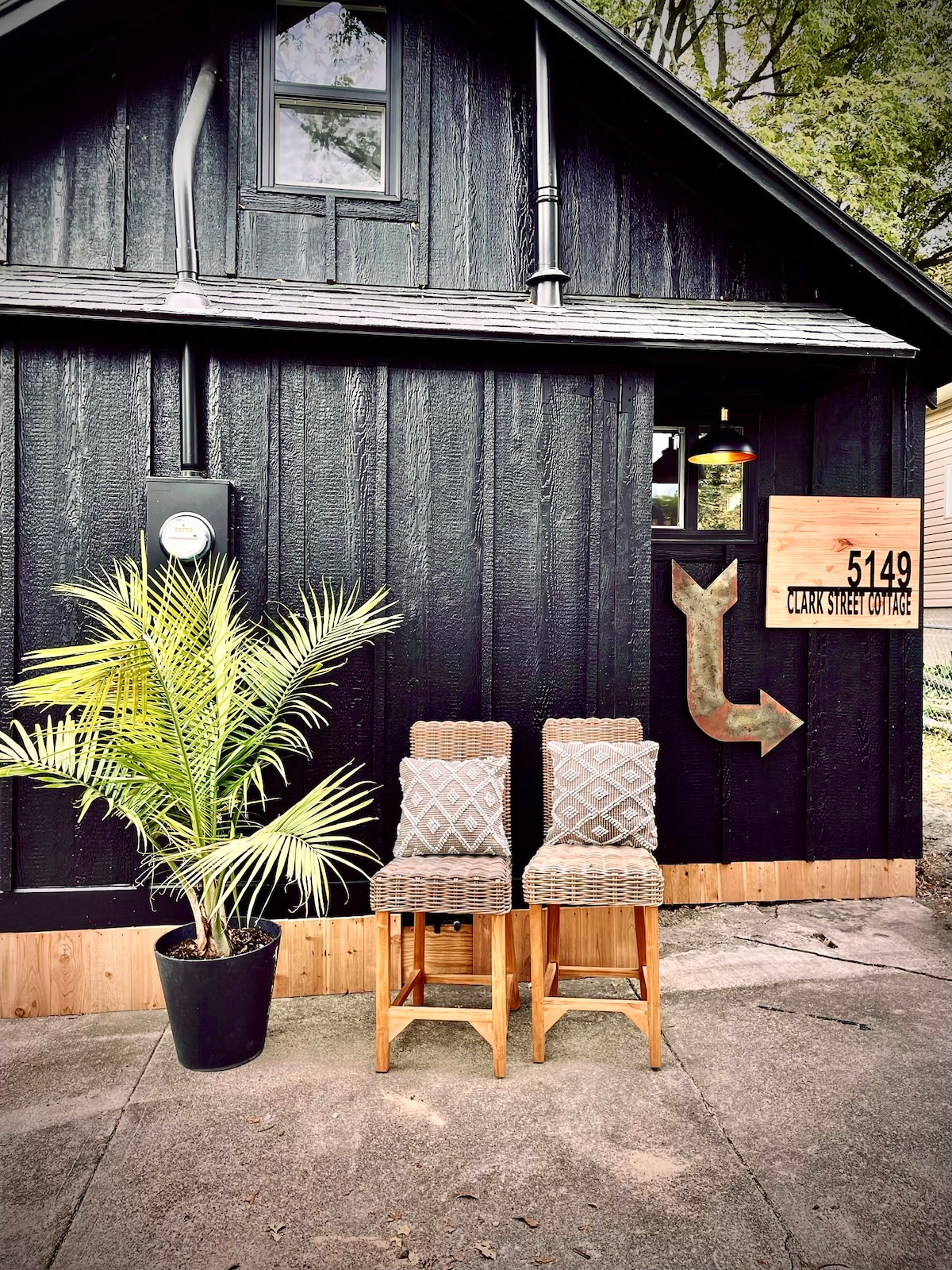 The exterior of Clark Street Cottage features a black wooden facade with wooden accents. Two tall bar stools are positioned beside a potted palm plant, creating a welcoming entrance. A wooden sign displays the address, while an arrow points towards the door.