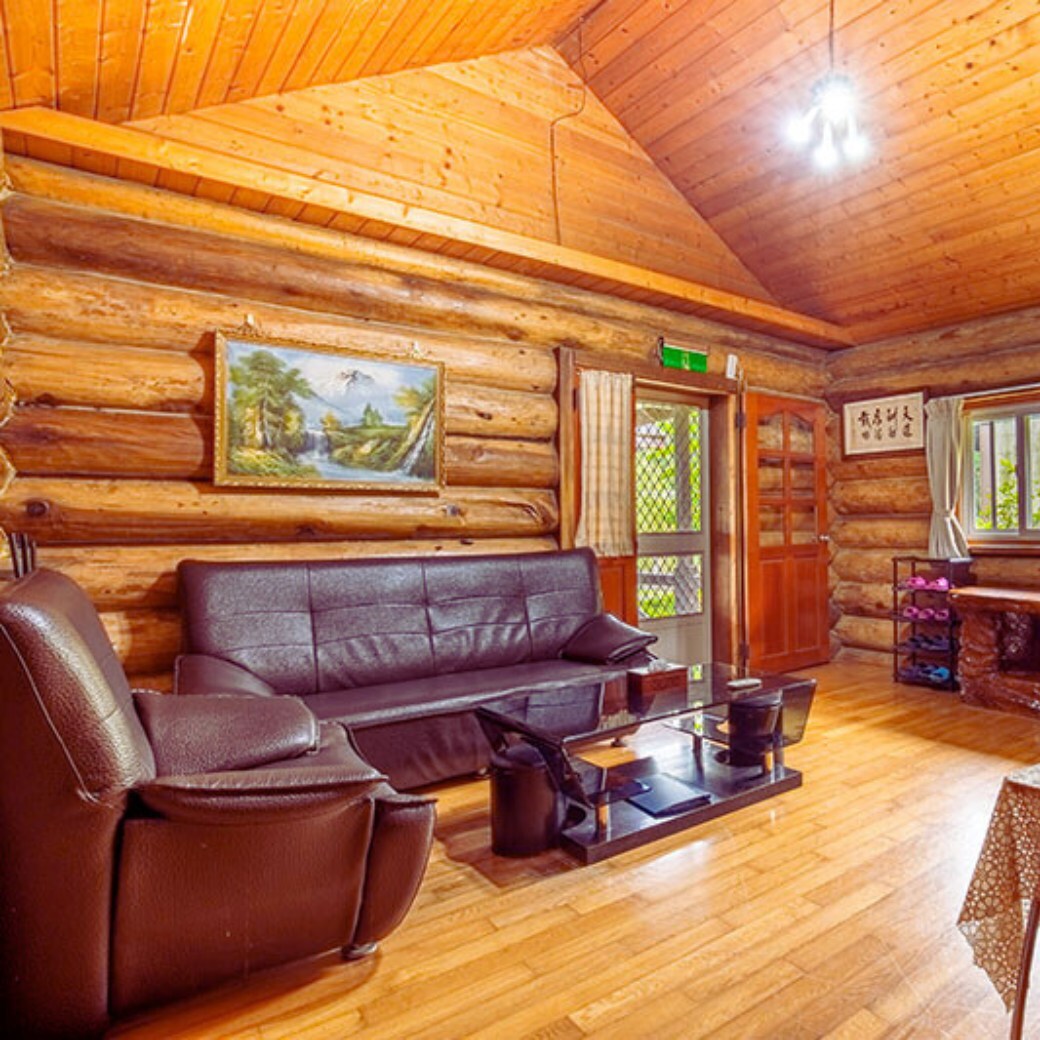 A spacious living area features logs as the main architectural element. A dark leather sofa and matching armchair are arranged around a low glass table. Natural light enters through the window, illuminating the warm wooden walls and ceiling.
