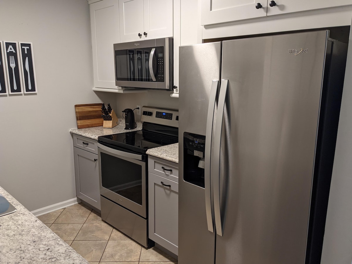 A modern kitchen is shown, featuring stainless steel appliances including a microwave, oven, and refrigerator. Light-colored cabinets are complemented by a granite countertop. A coffee maker and wooden cutting board are visible, contributing to the functional layout.