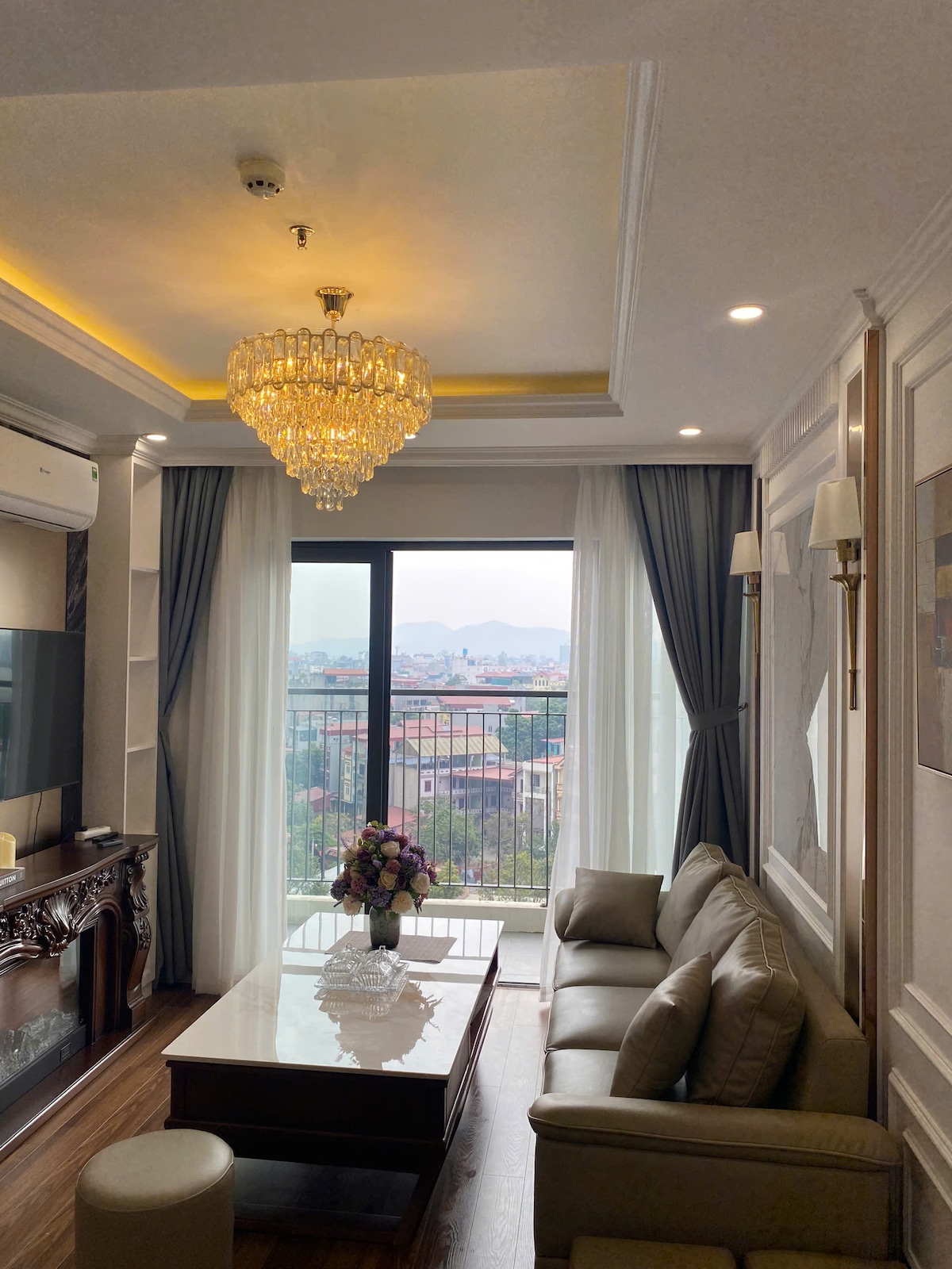 A modern living area features a stylish chandelier and a comfortable sofa arranged near a floor-to-ceiling window. Natural light filters in through the sheer curtains, illuminating the space. A decorative table sits at the center, adorned with a floral arrangement.