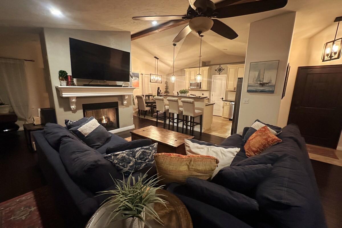 A spacious living area is showcased with a large sectional couch adorned with decorative pillows. A fireplace sits below a mounted television, and a wooden coffee table occupies the center. The kitchen is visible in the background, featuring modern appliances and counter seating.