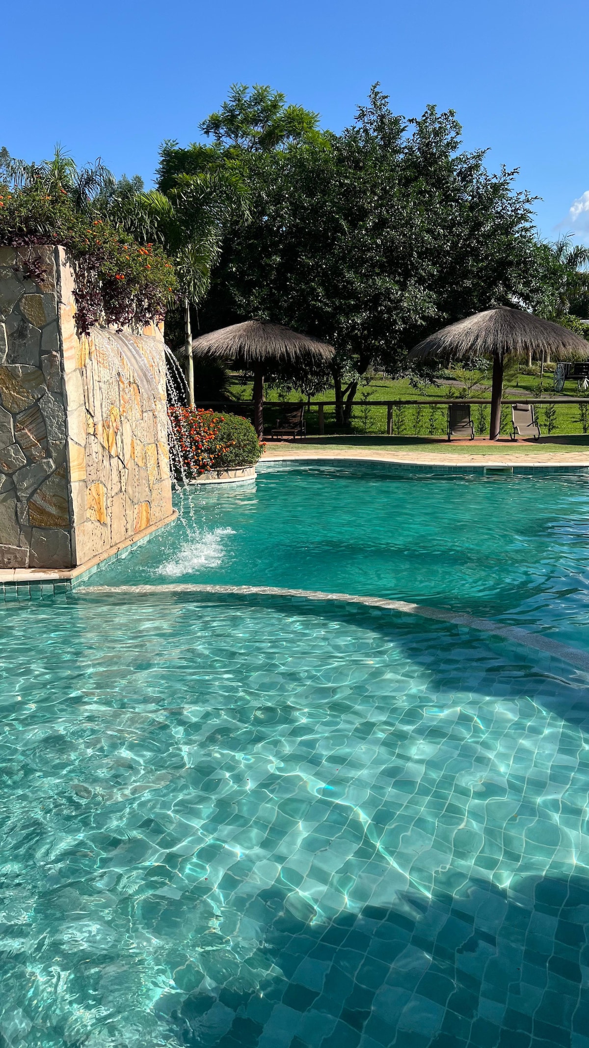 The inviting swimming pool features clear turquoise water, complemented by a cascading waterfall from a stone wall. Shaded seating areas with thatched roofs and surrounding lush greenery create a serene outdoor environment for relaxation.