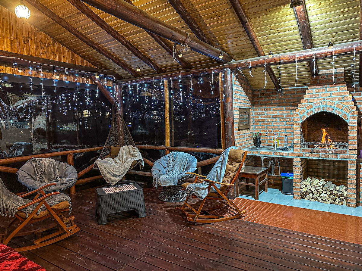 A cozy outdoor terrace is furnished with wooden rocking chairs and a small table, creating a relaxing space. A brick barbecue area is visible in the background, accompanied by neatly stacked firewood. Soft lighting and decorative string lights enhance the atmosphere.