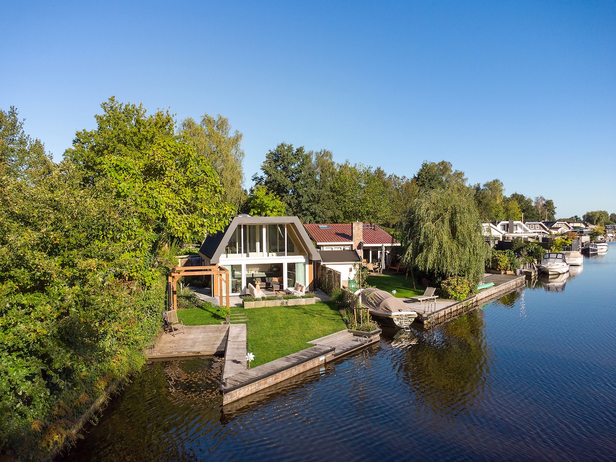 A modern structure with large glass panels is set beside a calm body of water, showcasing lush greenery and a neatly landscaped garden. A private dock is visible along the water's edge, while boats are moored nearby, providing easy access to the serene surroundings.