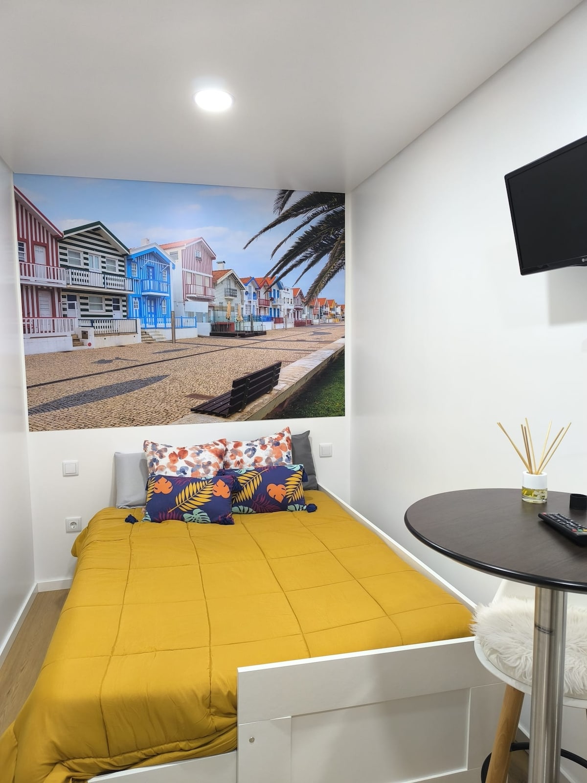 A small sleeping area features a yellow bedspread with decorative pillows in shades of blue and orange. A mural of colorful houses is displayed on the wall, evoking a scenic beach atmosphere. An adjacent round table with decorative items adds to the cozy ambiance.