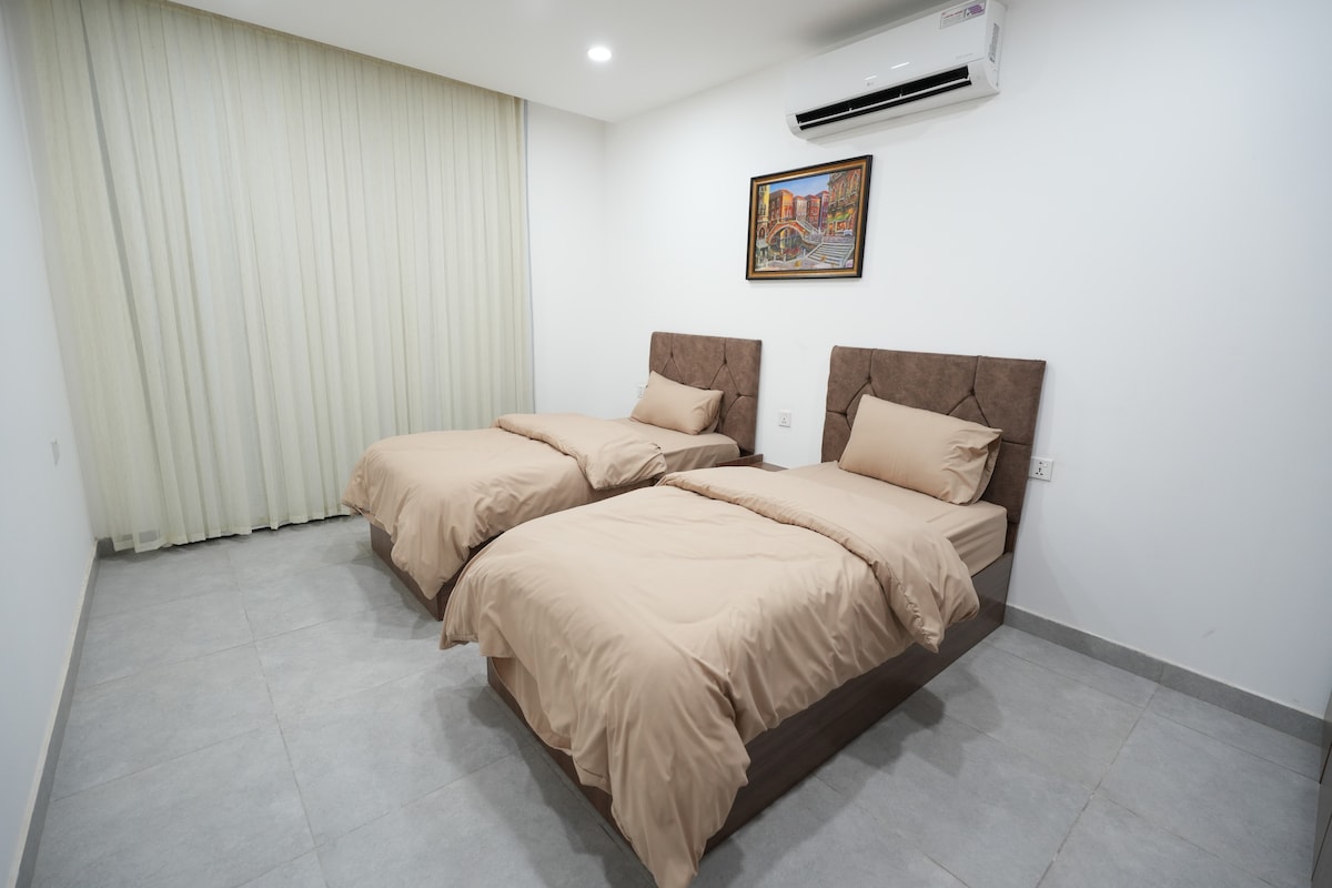 The sleeping area features two spacious beds with neutral-toned bedding. A piece of wall art is displayed above the headboards, and natural light filters through sheer curtains, enhancing the calm ambiance of the room.