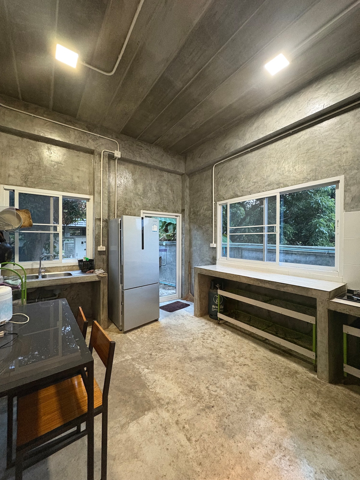 A functional kitchen features a concrete design with large windows providing natural light. A stainless steel refrigerator stands against the wall next to an entrance leading outside. A simple dining table with wooden chairs is positioned beside a spacious preparation area.