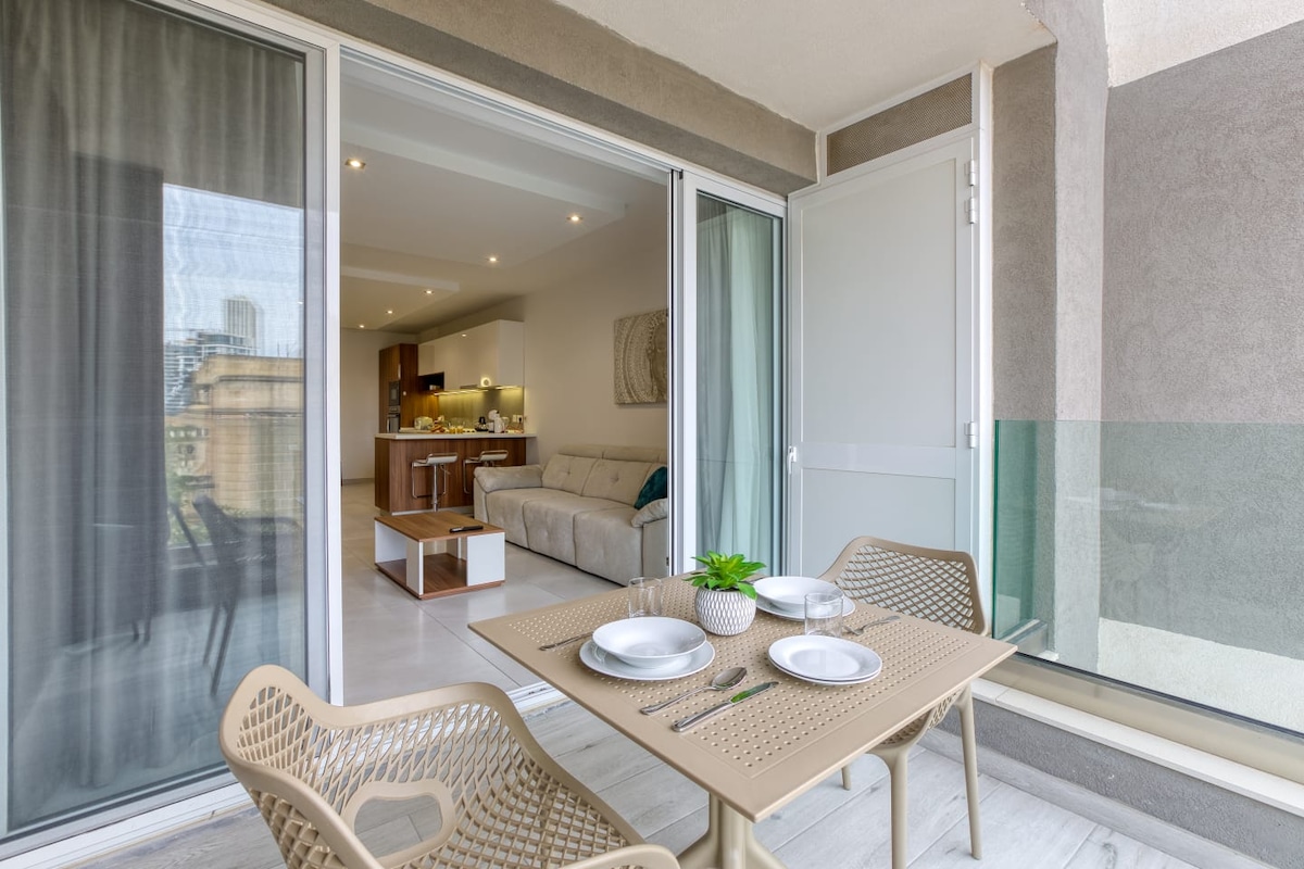 An outdoor terrace features a small dining table set for four, complemented by contemporary chairs. Glass sliding doors provide a view into a light-filled interior space, showcasing a comfortable sofa and an open-plan kitchen area.