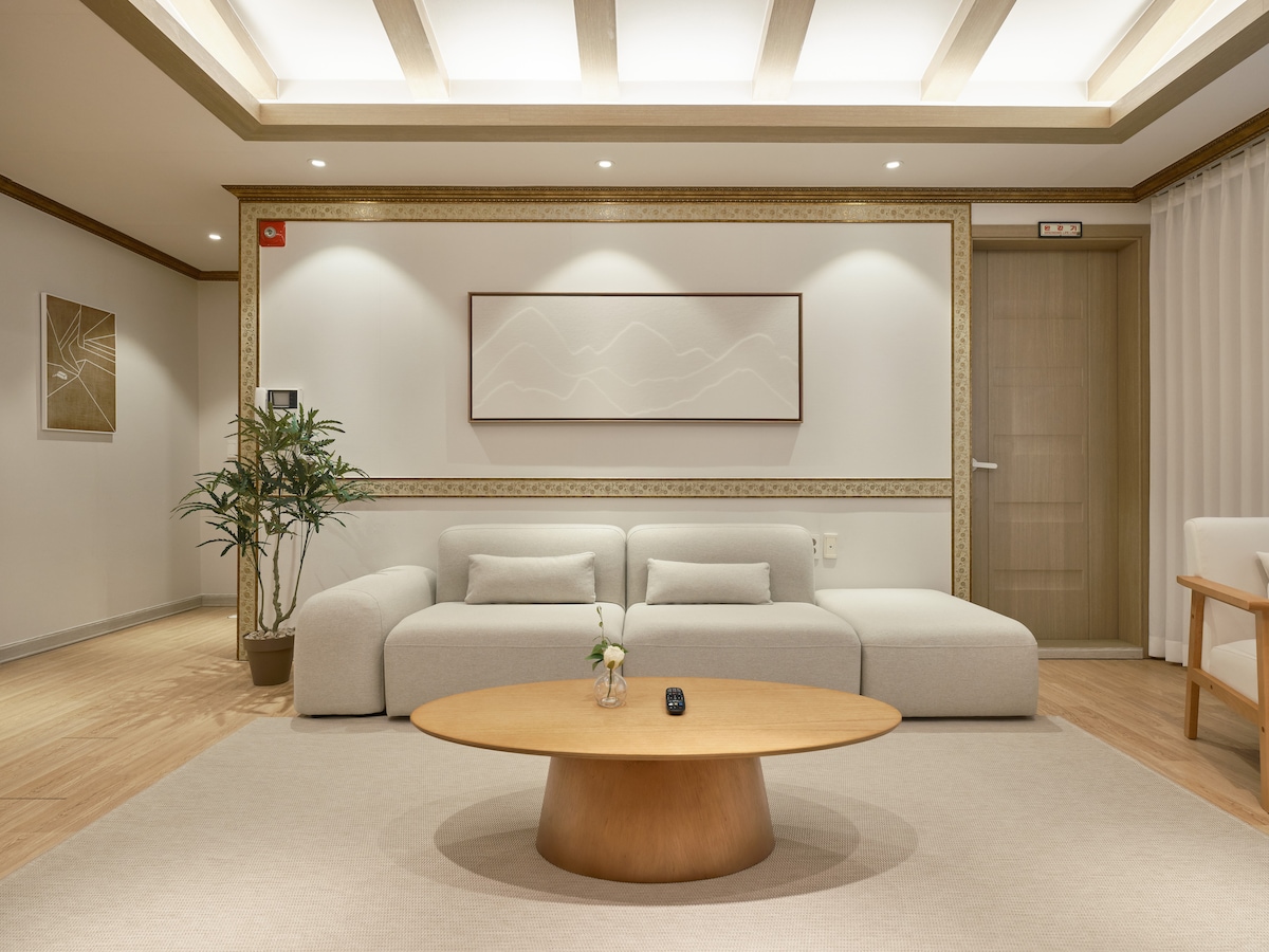 A spacious living area is featured with a soft, neutral color palette. A large circular wooden coffee table sits centrally, surrounded by a comfortable sectional sofa. Decorative plants add a touch of nature, while soft lighting enhances the inviting atmosphere.