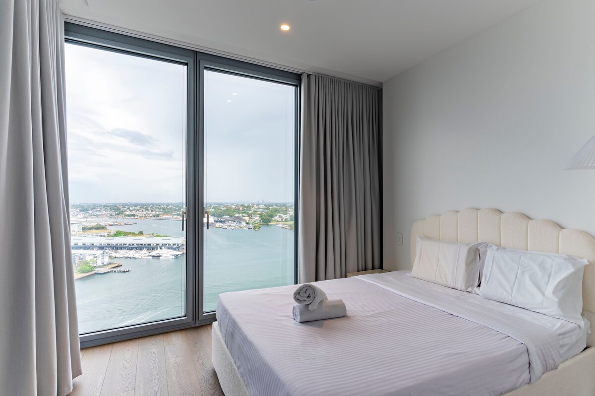 A serene bedroom features a spacious bed with crisp linens and a neatly rolled towel placed on top. Large windows dominate one side, offering sweeping water views and natural light. Neutral-toned curtains frame the windows, complementing the soft color palette of the room.